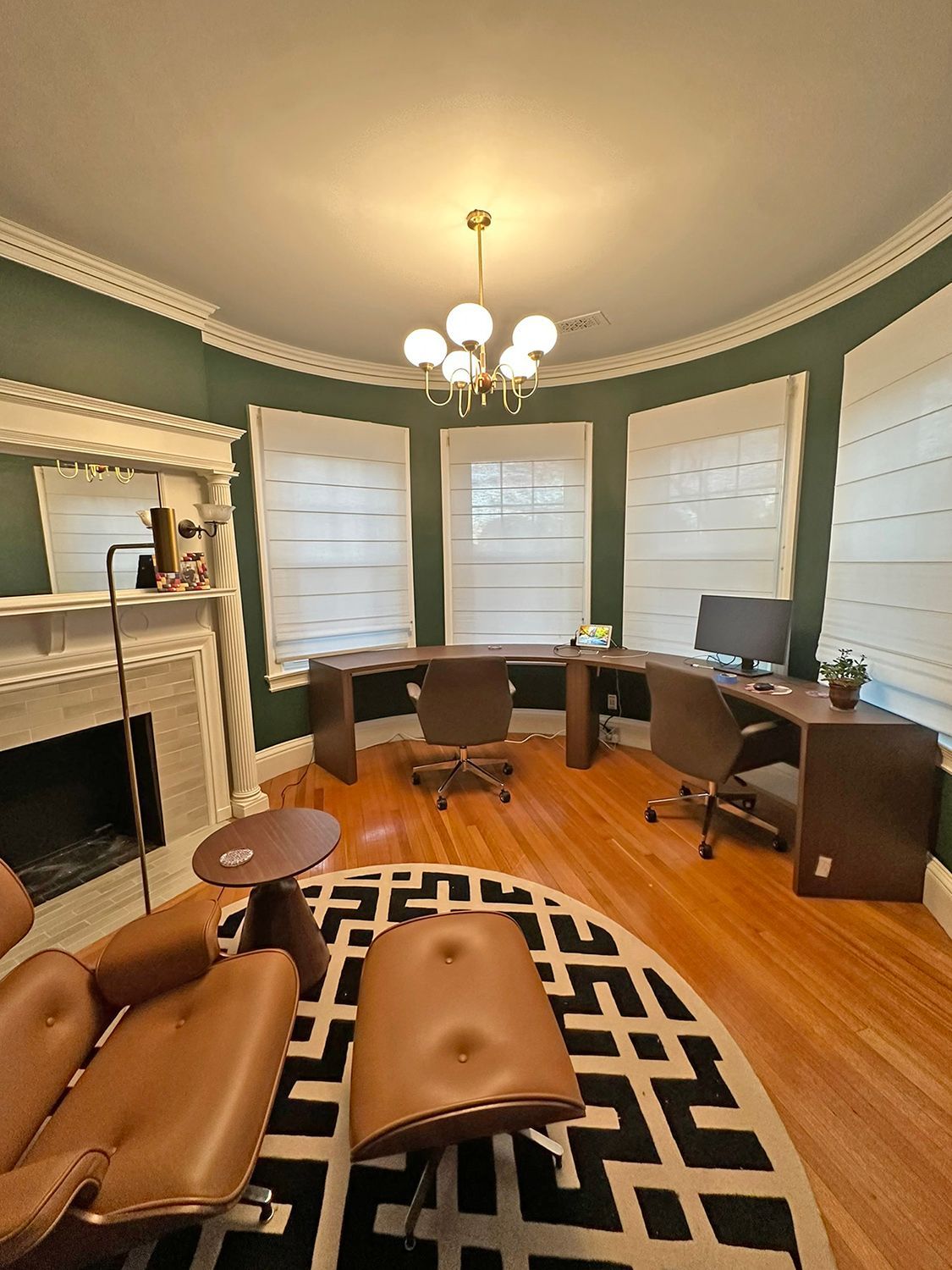 An office with a curved wall of windows, dark wood desk, and a leather chair and ottoman on a patterned rug.