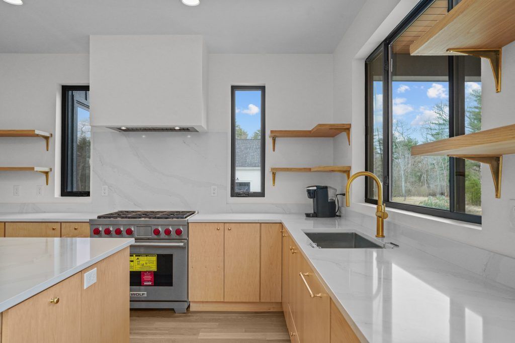 Modern kitchen with light wood cabinets, white countertops, stainless steel appliances, and large windows.