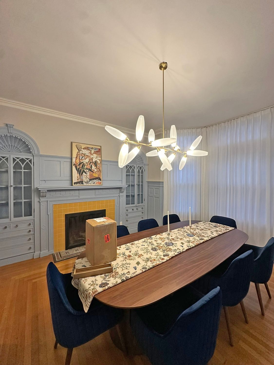 Dining room with a long wooden table, blue chairs, fireplace, and leaf-shaped chandelier.