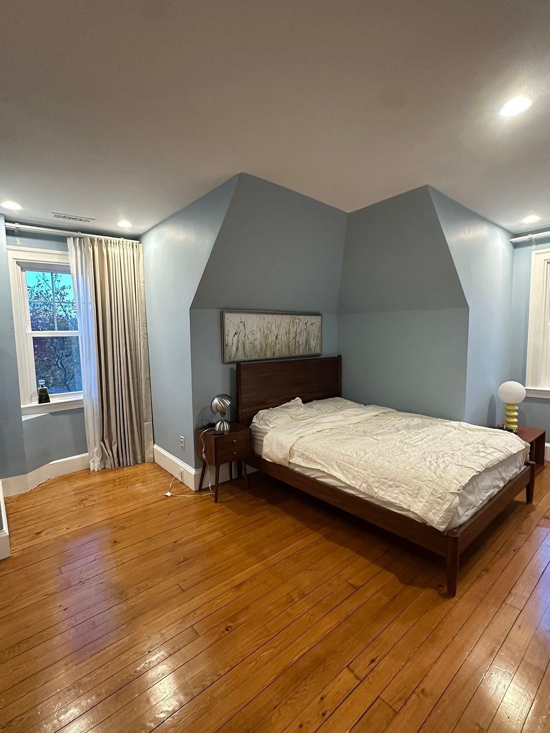 Bedroom with light blue walls, wood bed, and hardwood floors. White bedding, curtains, and two windows.