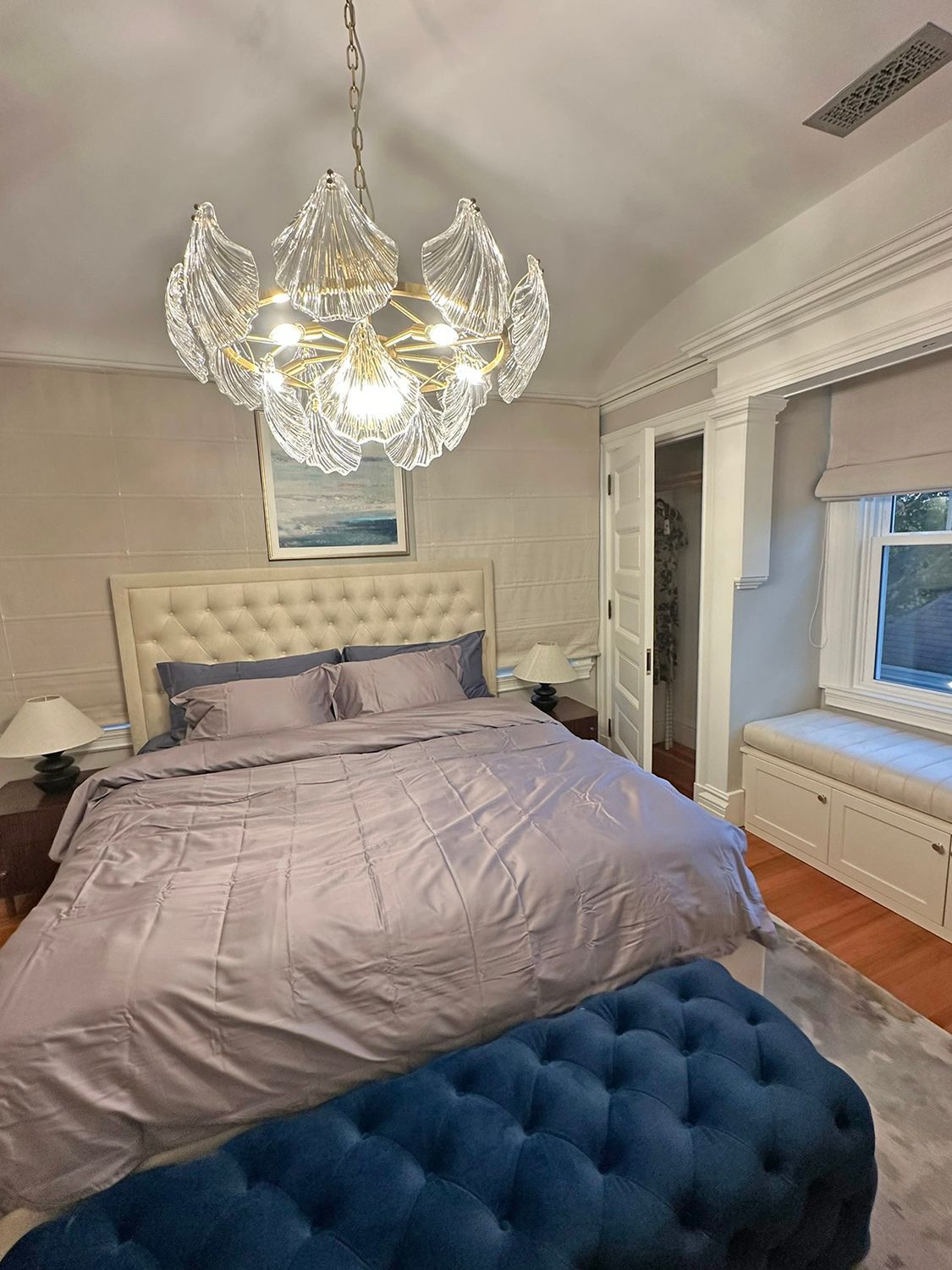 Bedroom with a crystal chandelier, tufted bed, and velvet ottoman. Soft lilac bedding. White walls and window seat.