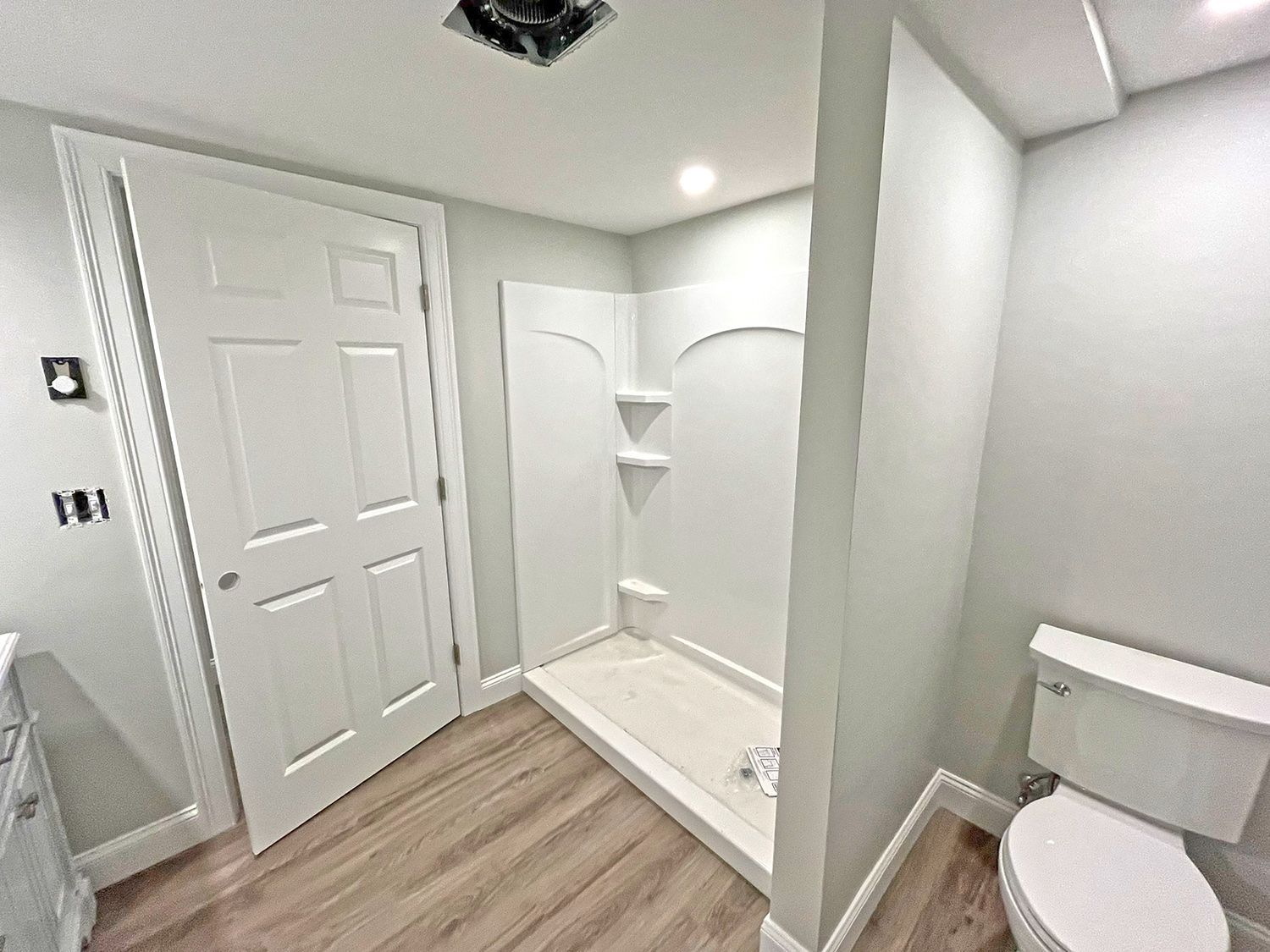 Bathroom with white door, shower stall, toilet, and gray walls, and light-colored wood-look flooring.