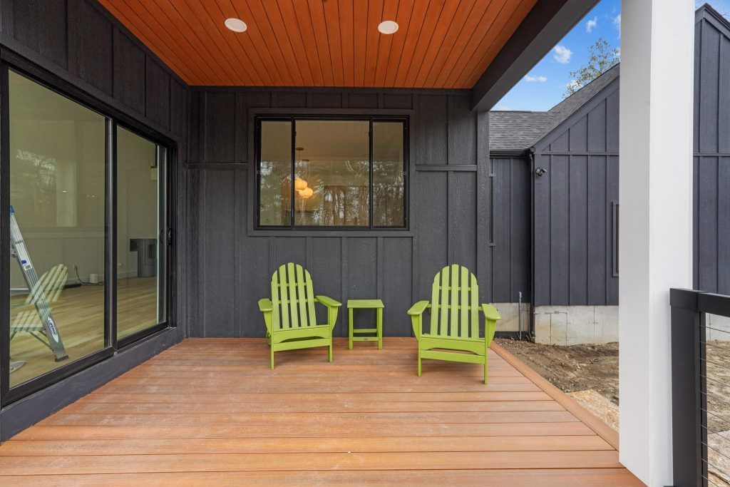 A covered porch with two green Adirondack chairs and a small table. Black siding and wood deck.