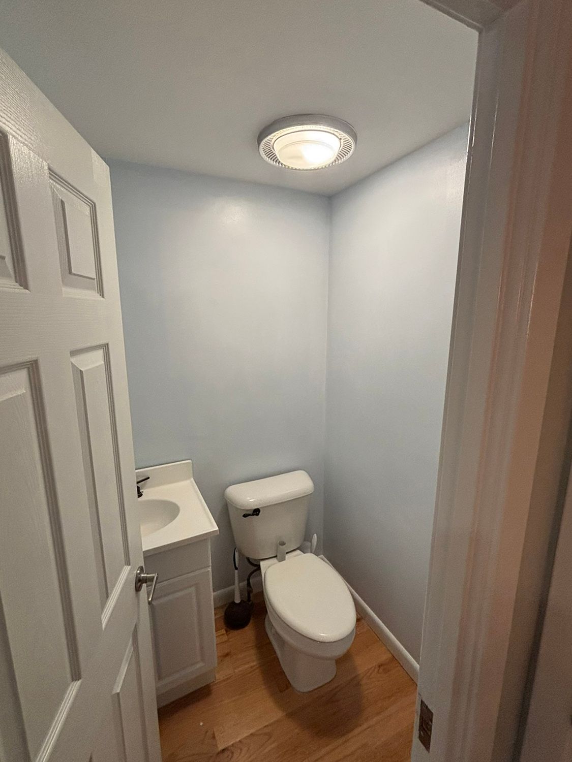 Small bathroom with toilet, sink, and closed white door. Light blue walls, wood floor.