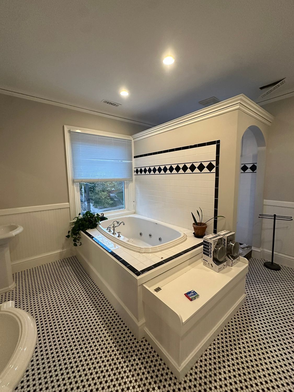 Bathroom with a white jacuzzi tub, black and white tiled floor, and a window with a blind.