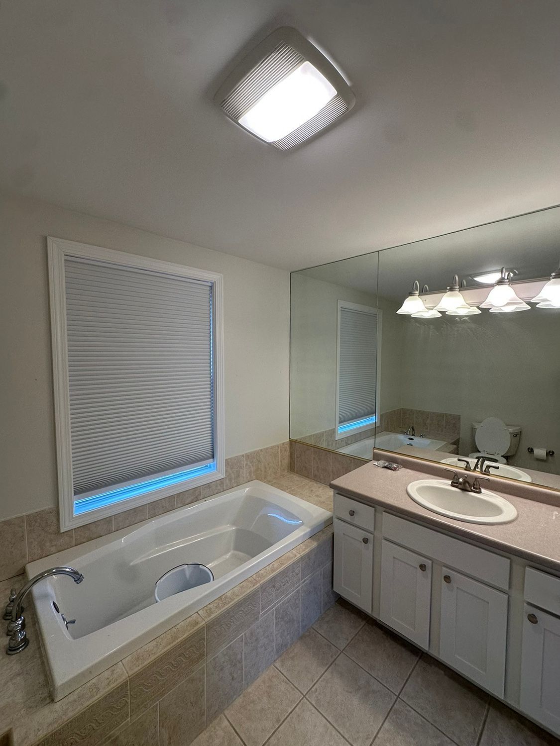 Bathroom with a white bathtub, vanity, and large mirror. Window with closed shade. Beige tile.