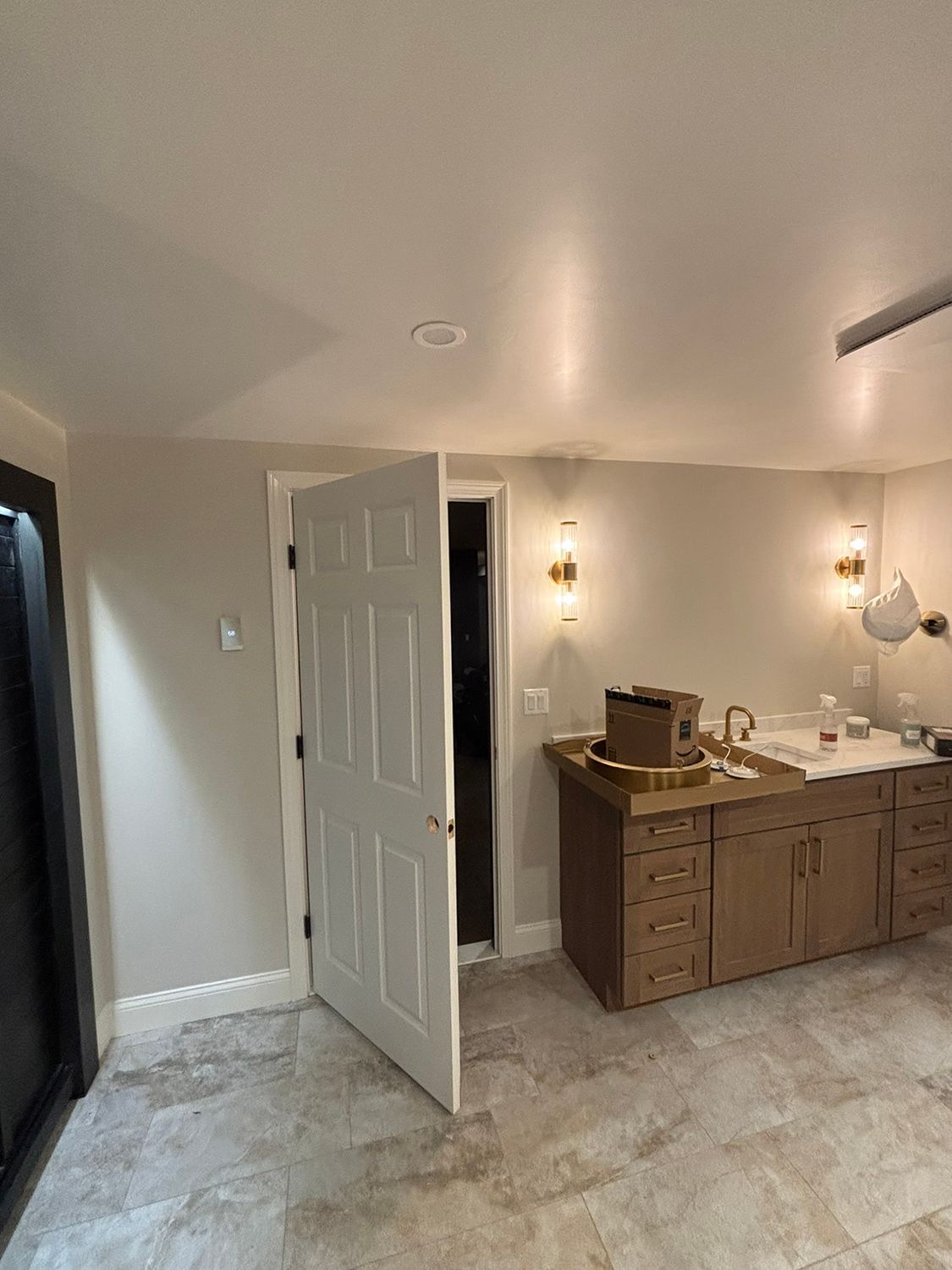 Bathroom with open white door, brown vanity, gold sconces, and light-colored tiled floor.