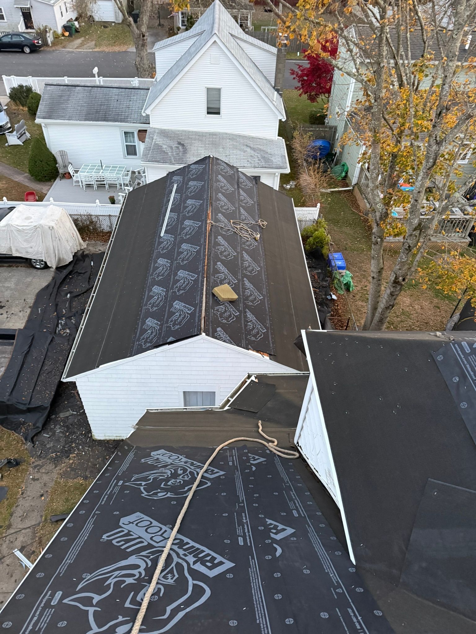 Overhead view of a roof under construction; black roofing material laid down, surrounding houses, some with autumn foliage.