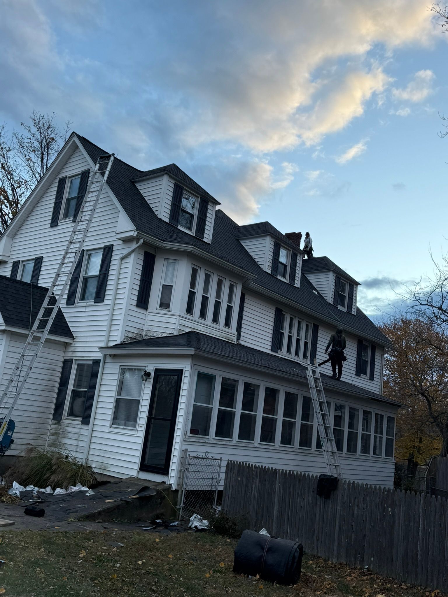 White multi-story house with black roof, workers on ladders. Cloudy sky background.
