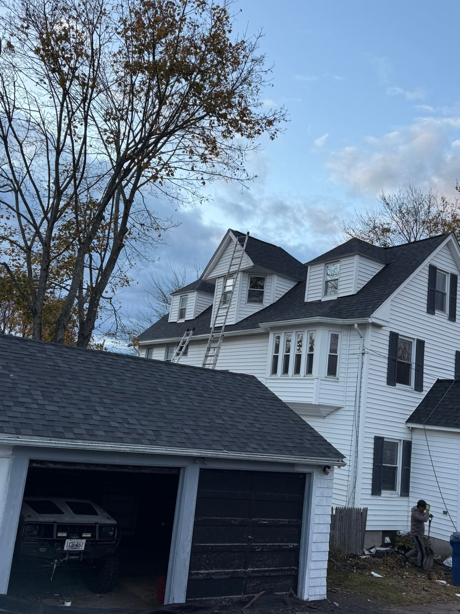 White house with dark roof and attached garage; ladder against upper story under overcast sky.