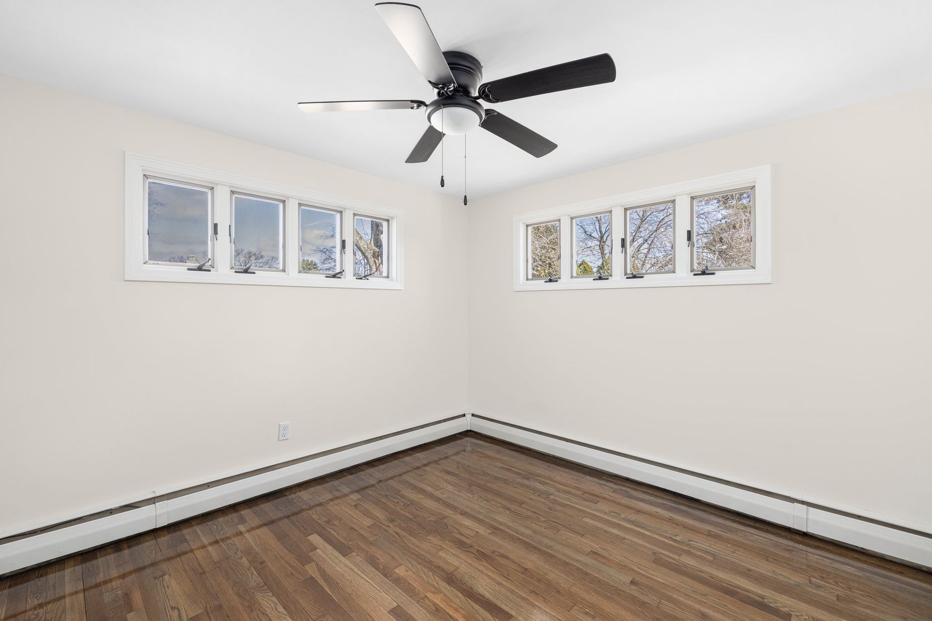 Empty room with hardwood floor, two windows, and a ceiling fan. Walls are cream colored.