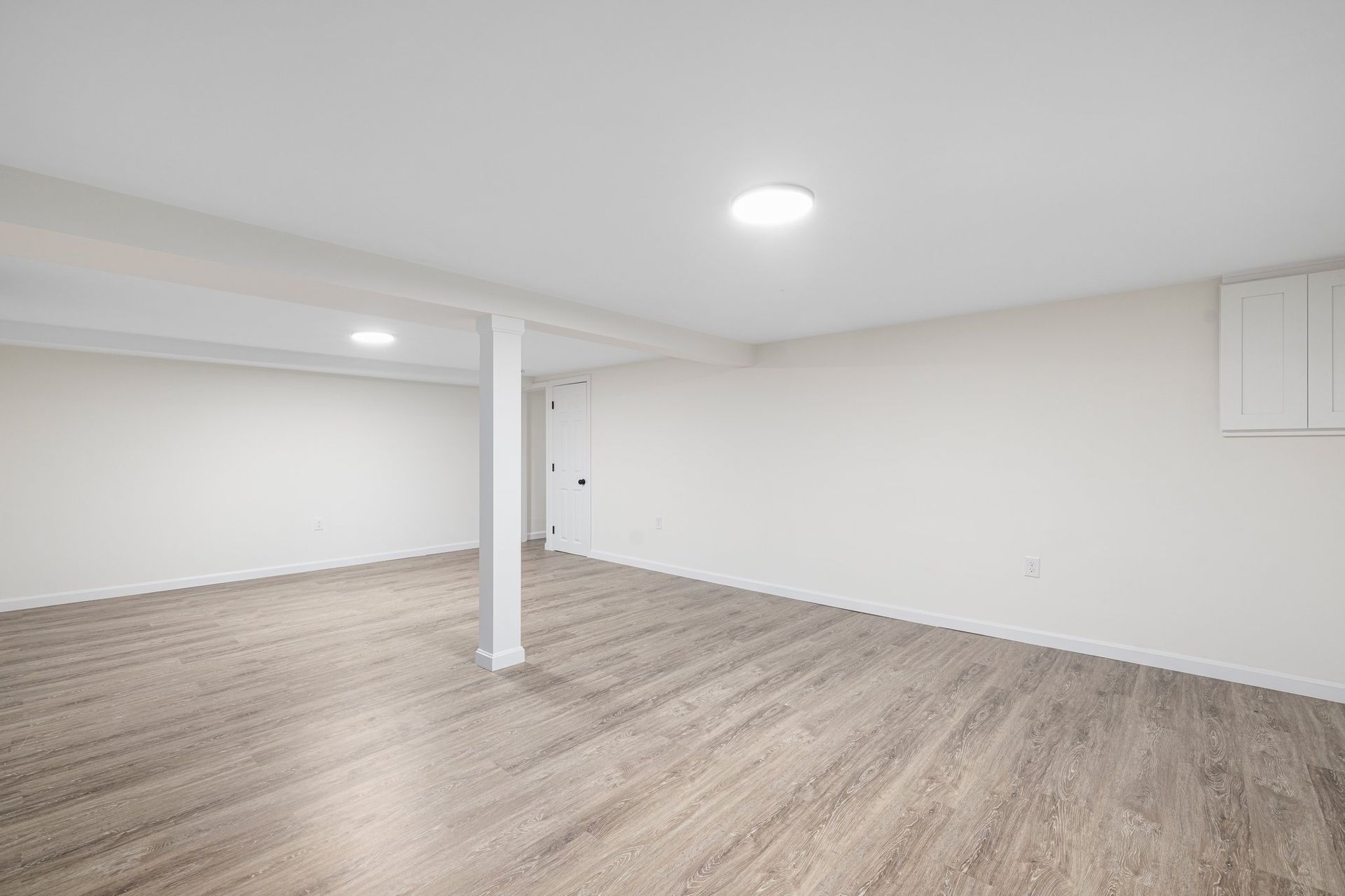 Empty room with light wood-look flooring, white walls, ceiling lights, and support beams.
