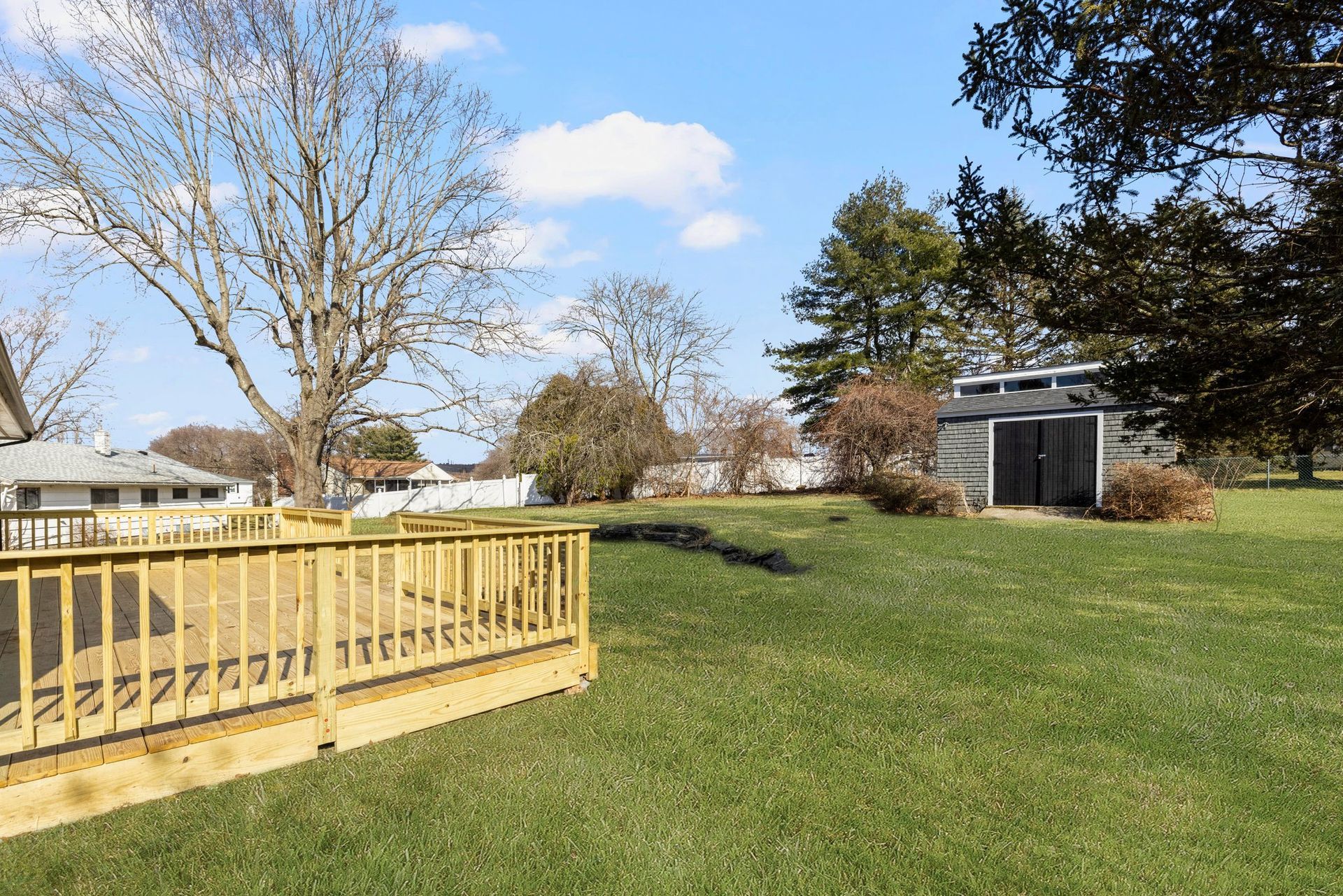Backyard with a wooden deck, green grass, trees, and a gray shed. Bright, sunny day.