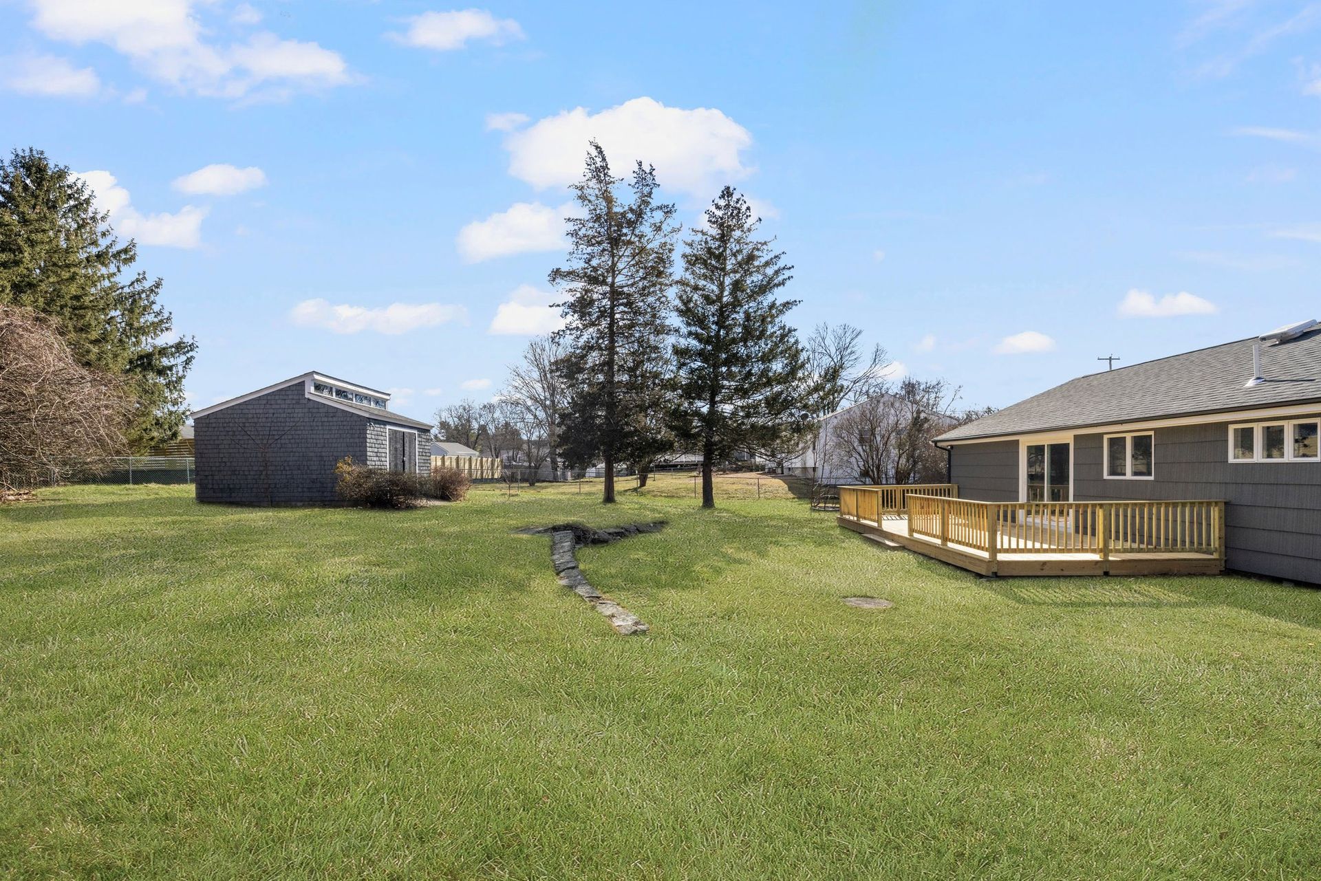 Grassy backyard with deck, shed, and trees under a blue sky.