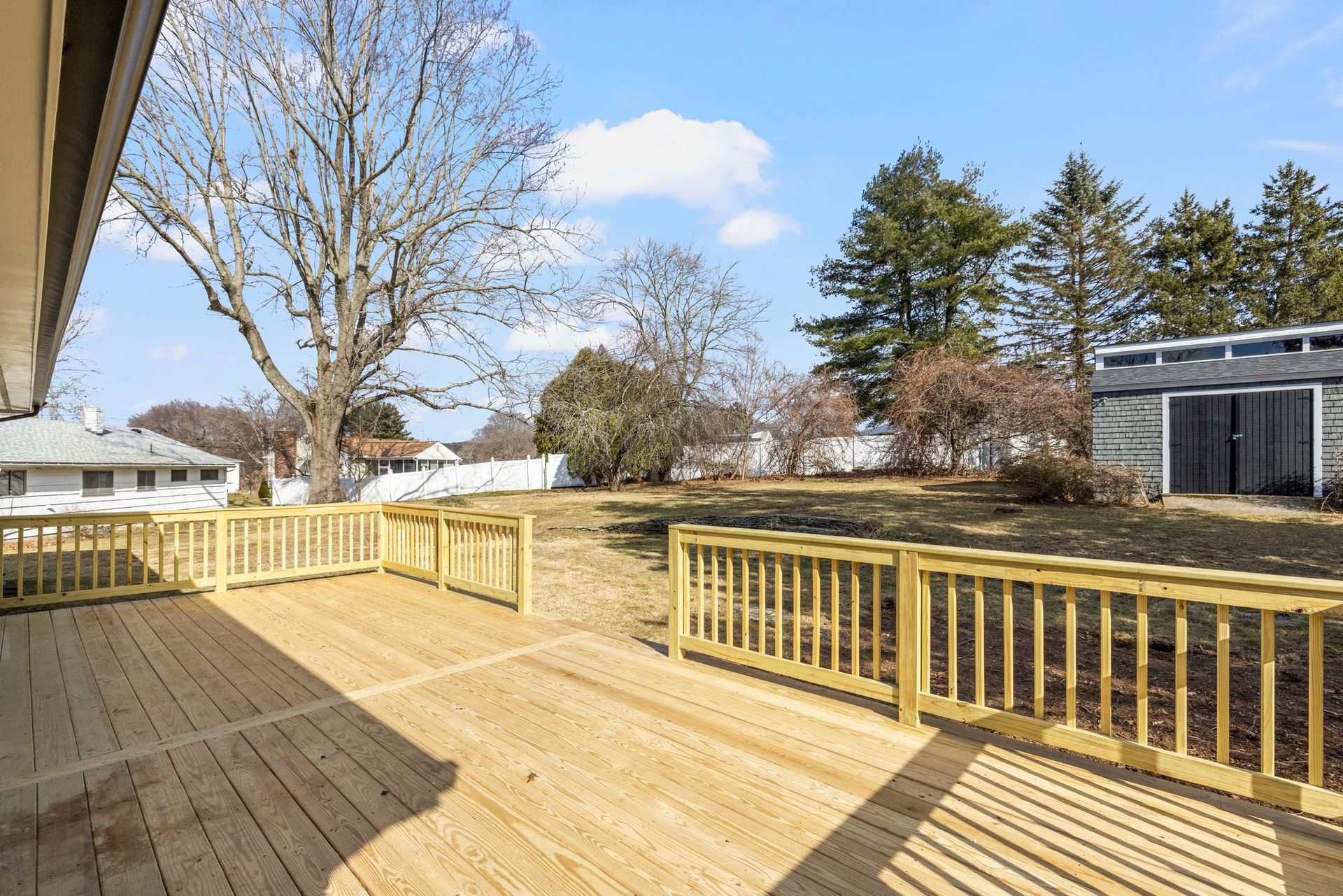 Wooden deck in a backyard with a shed, trees, and blue sky.