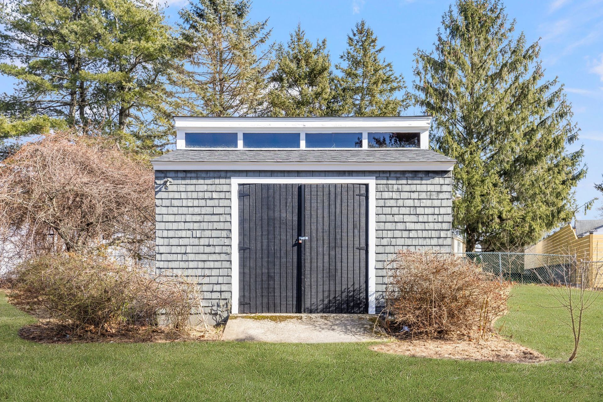 Gray shed with black doors, small windows, on a green lawn with bushes and trees in the background.