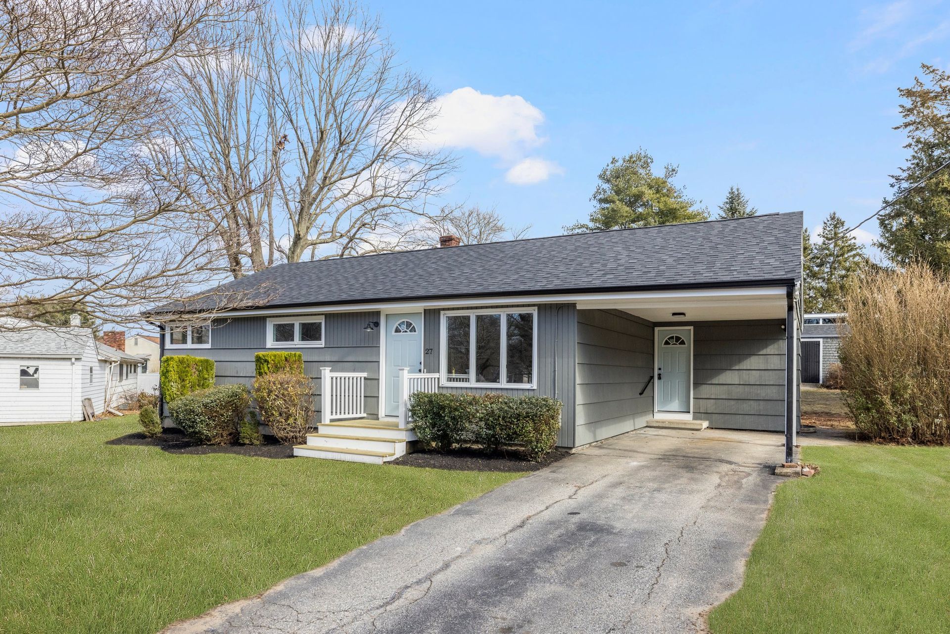 Gray ranch-style house with carport and driveway on a green lawn; cloudy sky.