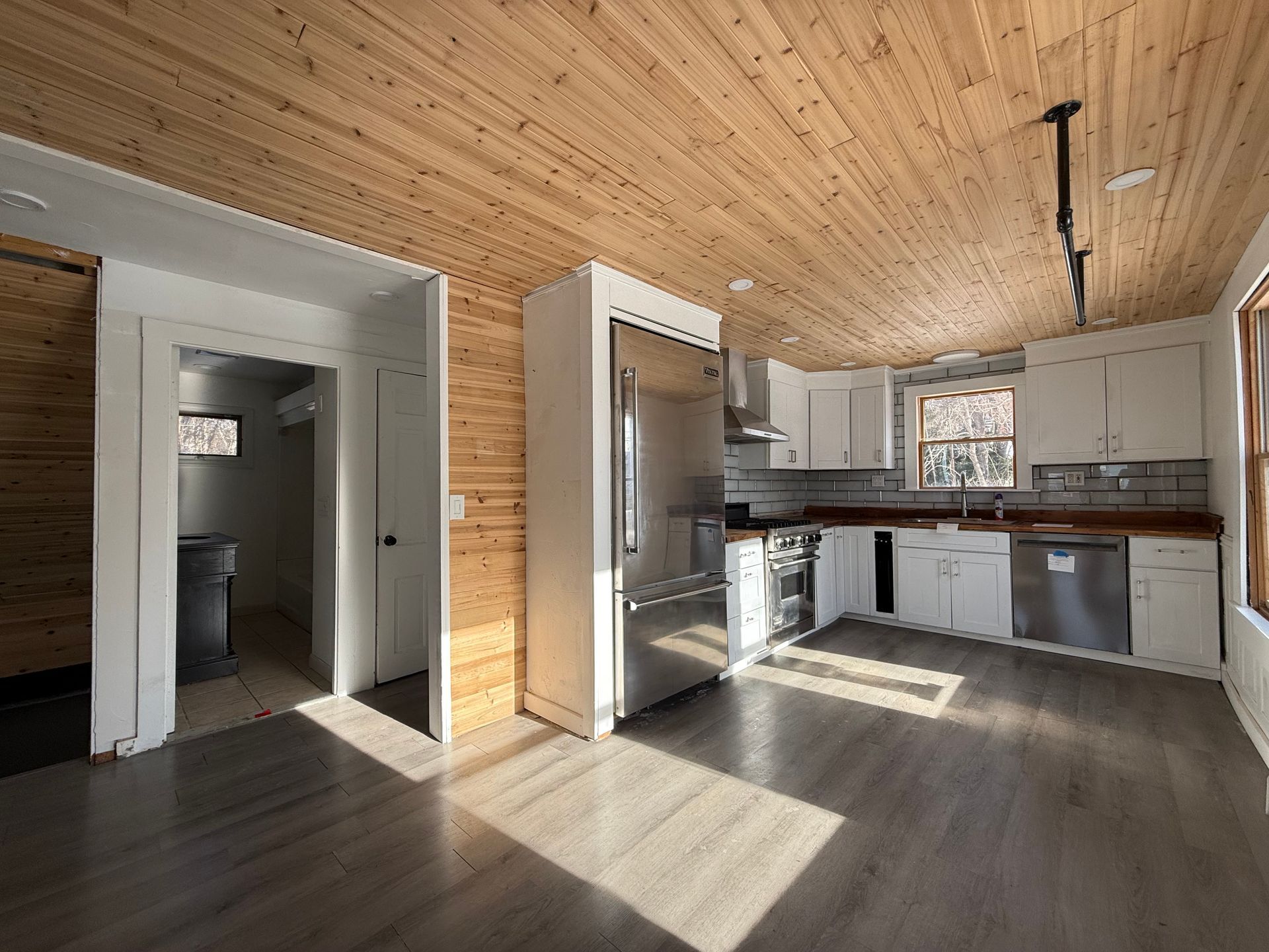 Kitchen with wood ceiling and floors. Stainless steel appliances, white cabinets, and a doorway to a bathroom.