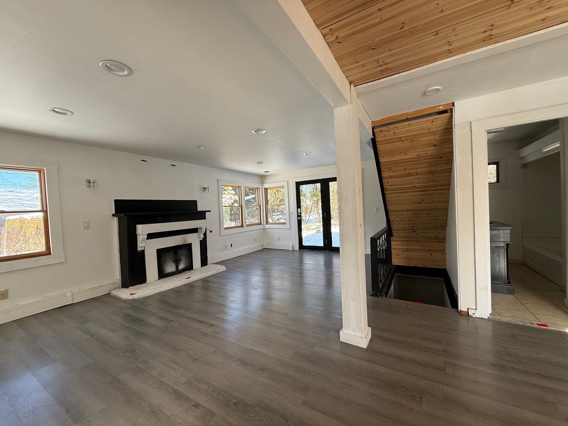 Empty living room with fireplace, windows, hardwood floor, and a wooden ceiling.