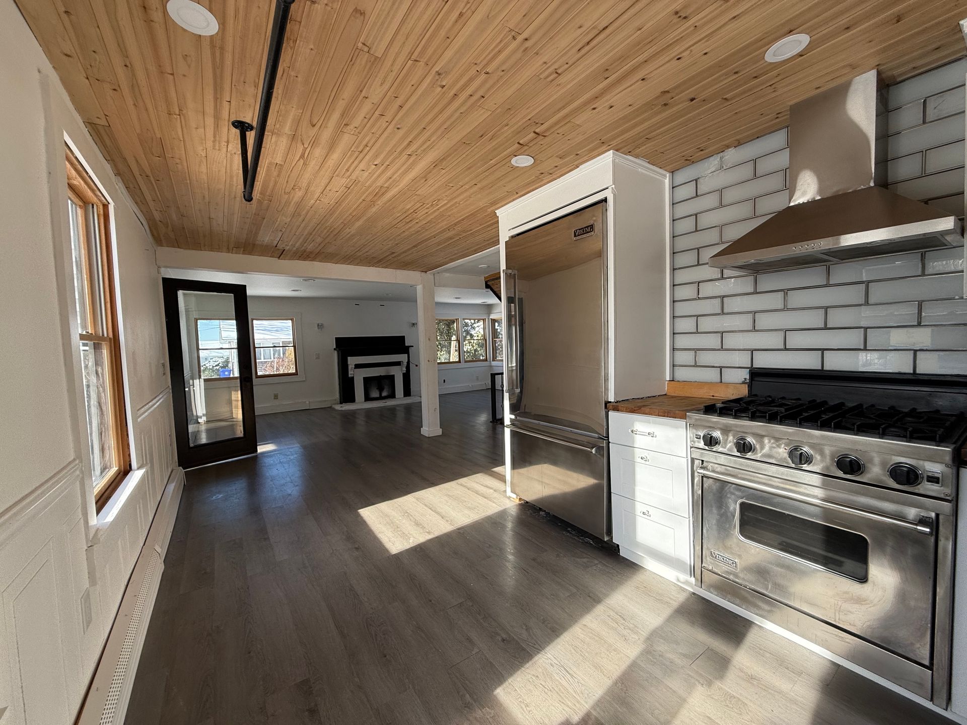Modern kitchen with stainless steel appliances, wood ceiling, and exposed brick backsplash, adjacent to a living area.