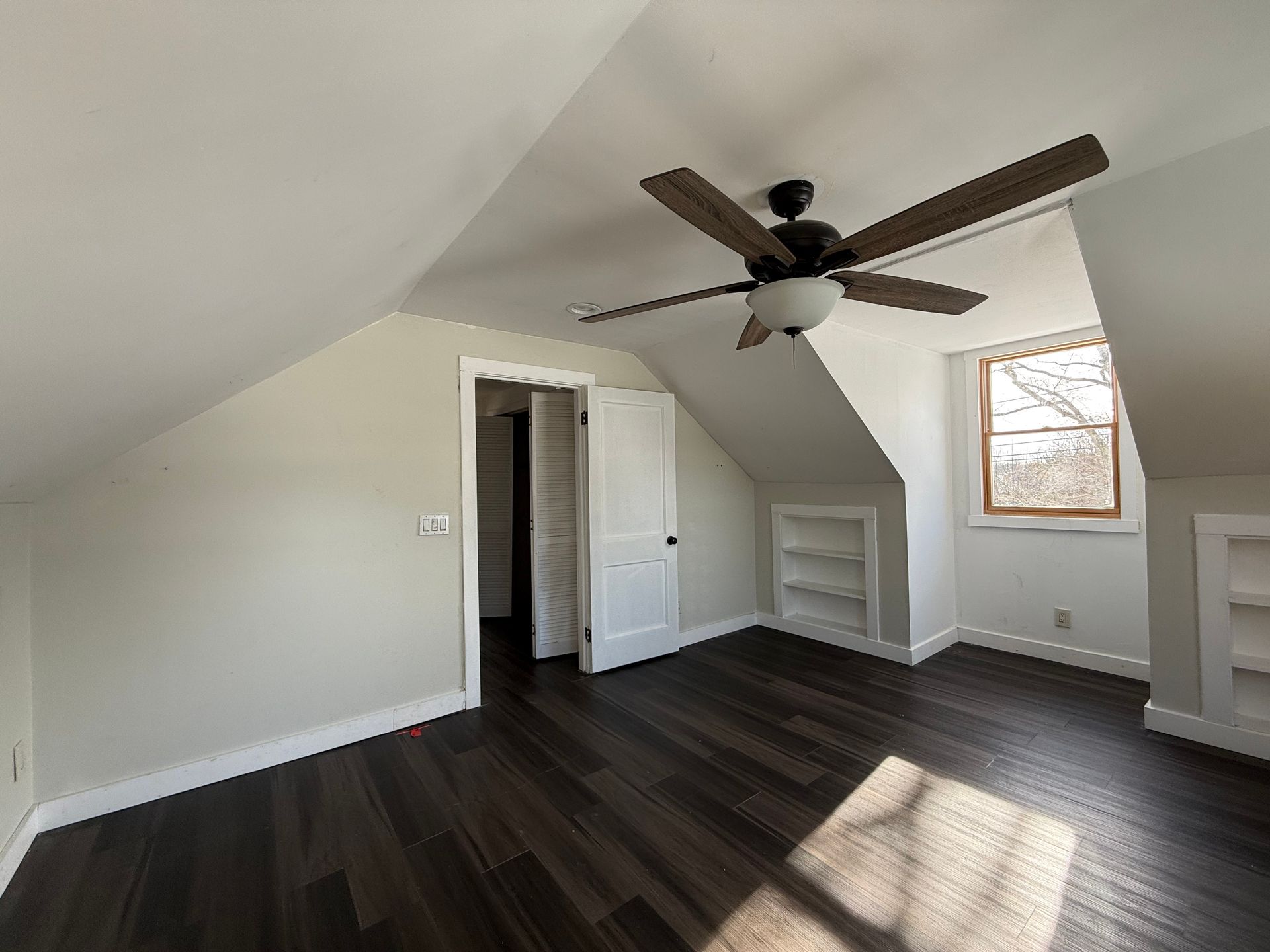 Attic bedroom with dark wood floor, white walls, built-in shelving, and a ceiling fan.