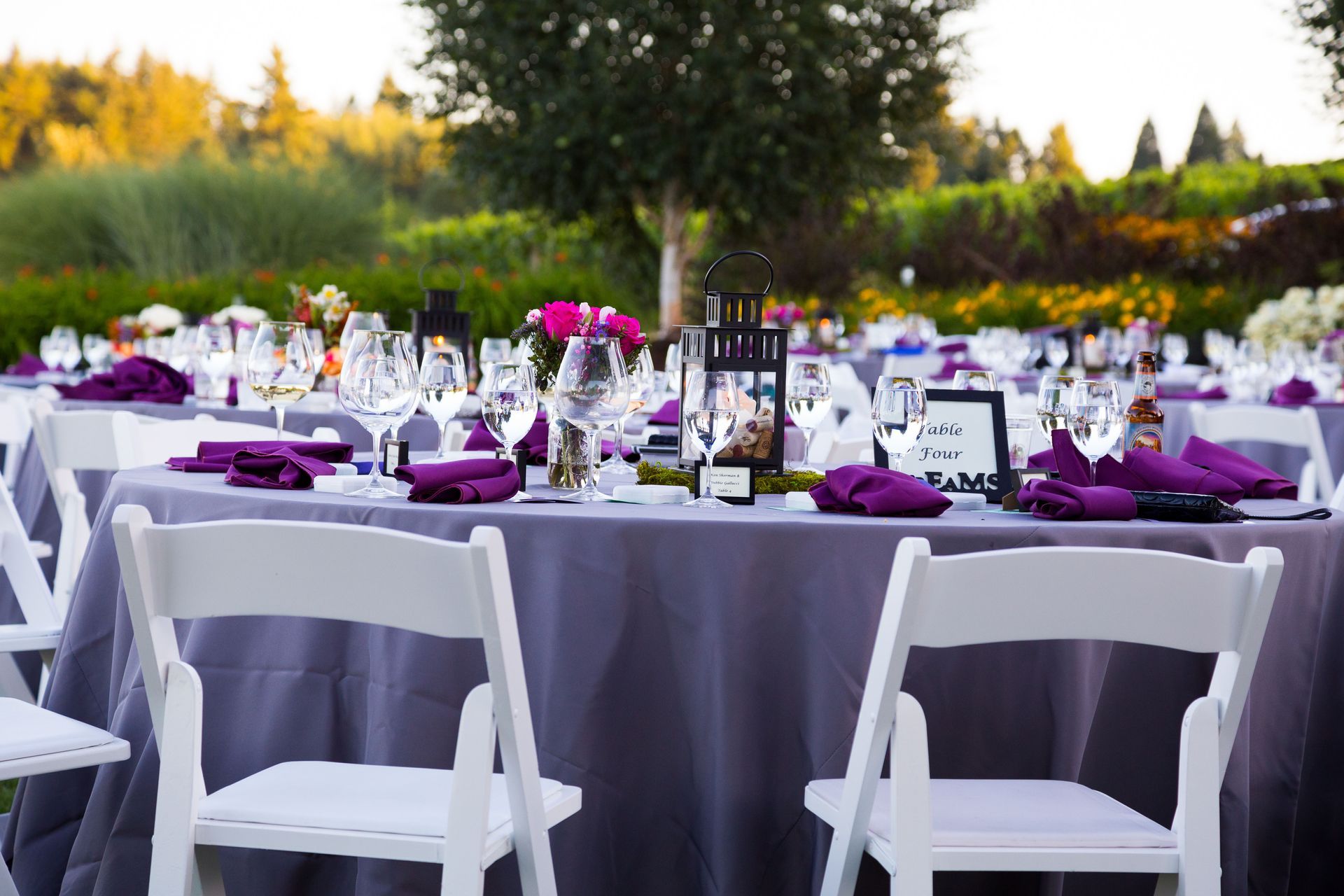 Outdoor reception tables set with purple linens and napkins, white chairs, and floral centerpieces.