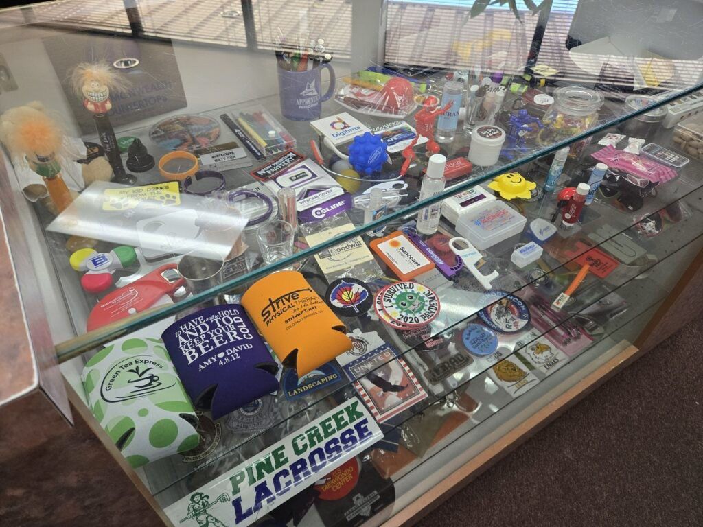 A sampling of various types of promotional items that Shirt Stop customizes, including koozies, lip balms, patches, and more. (Photo by Breeanna Jent/The Gazette)