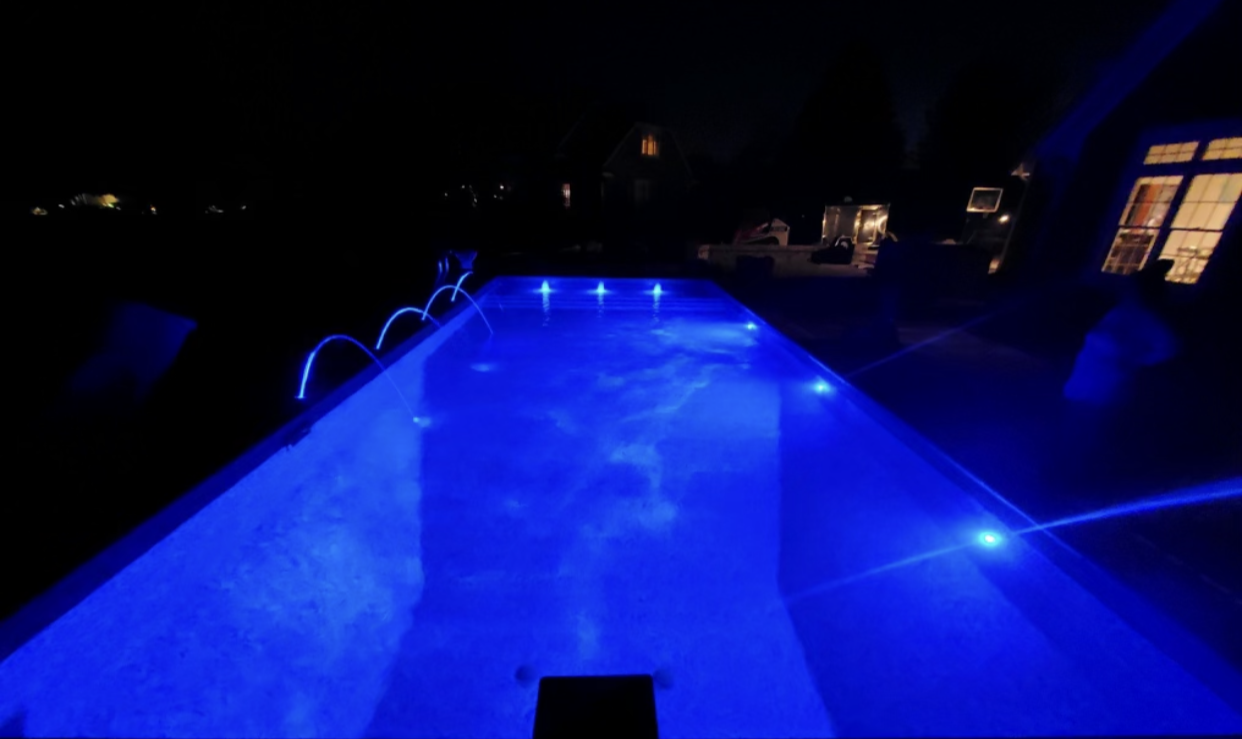 Nighttime view of a rectangular pool with blue lights illuminating the water, trees and a house are in the background.
