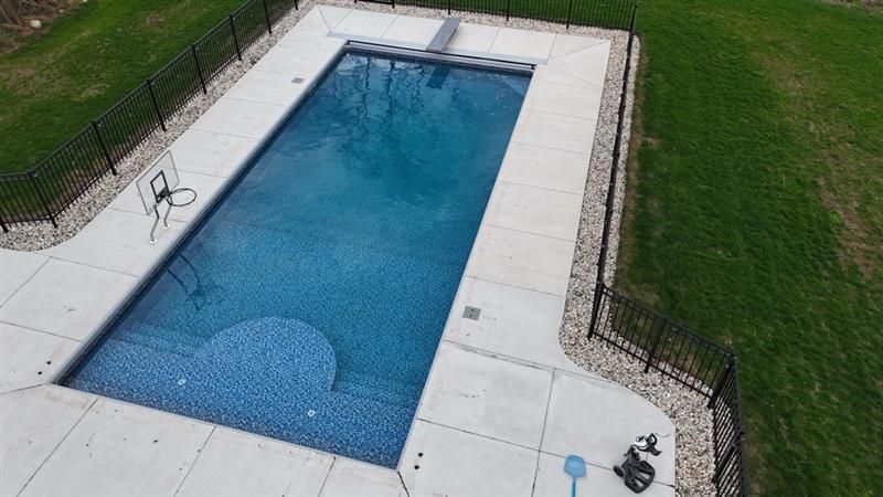 Aerial view of a rectangular blue tiled swimming pool with a diving board and basketball hoop, surrounded by concrete and grass.