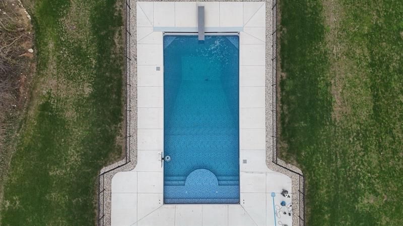 Overhead view of a rectangular blue swimming pool with a diving board, surrounded by concrete and grass.
