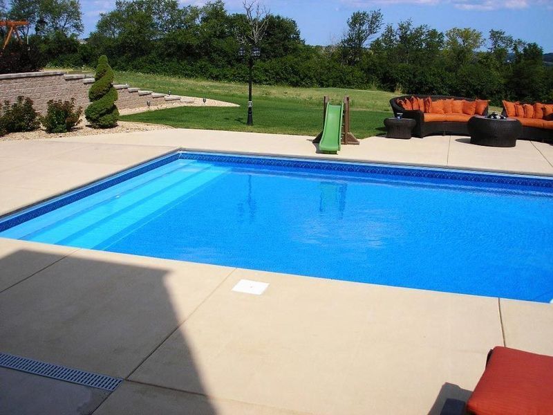 Outdoor rectangular swimming pool with concrete deck and lounge furniture, sunny day.