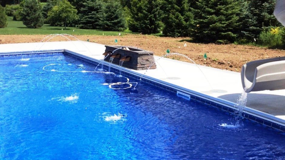 A blue swimming pool with fountains spraying water, concrete edging, and a slide; outdoors.