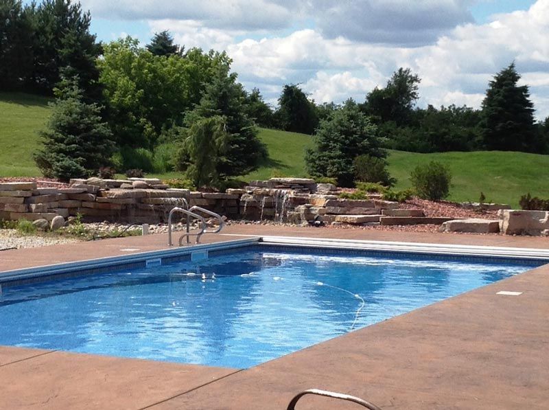 Swimming pool with blue water and brown concrete deck, overlooking a grassy hill and trees.