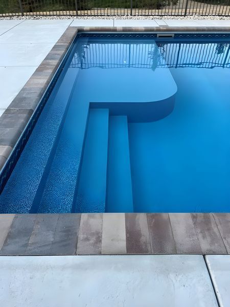 Swimming pool with built-in steps and a curved section. Blue water, gray tile edging, white concrete.