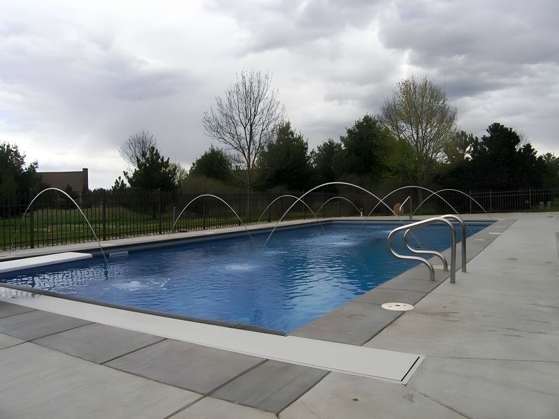 Rectangular blue swimming pool with fountain jets, surrounded by concrete and a cloudy sky.