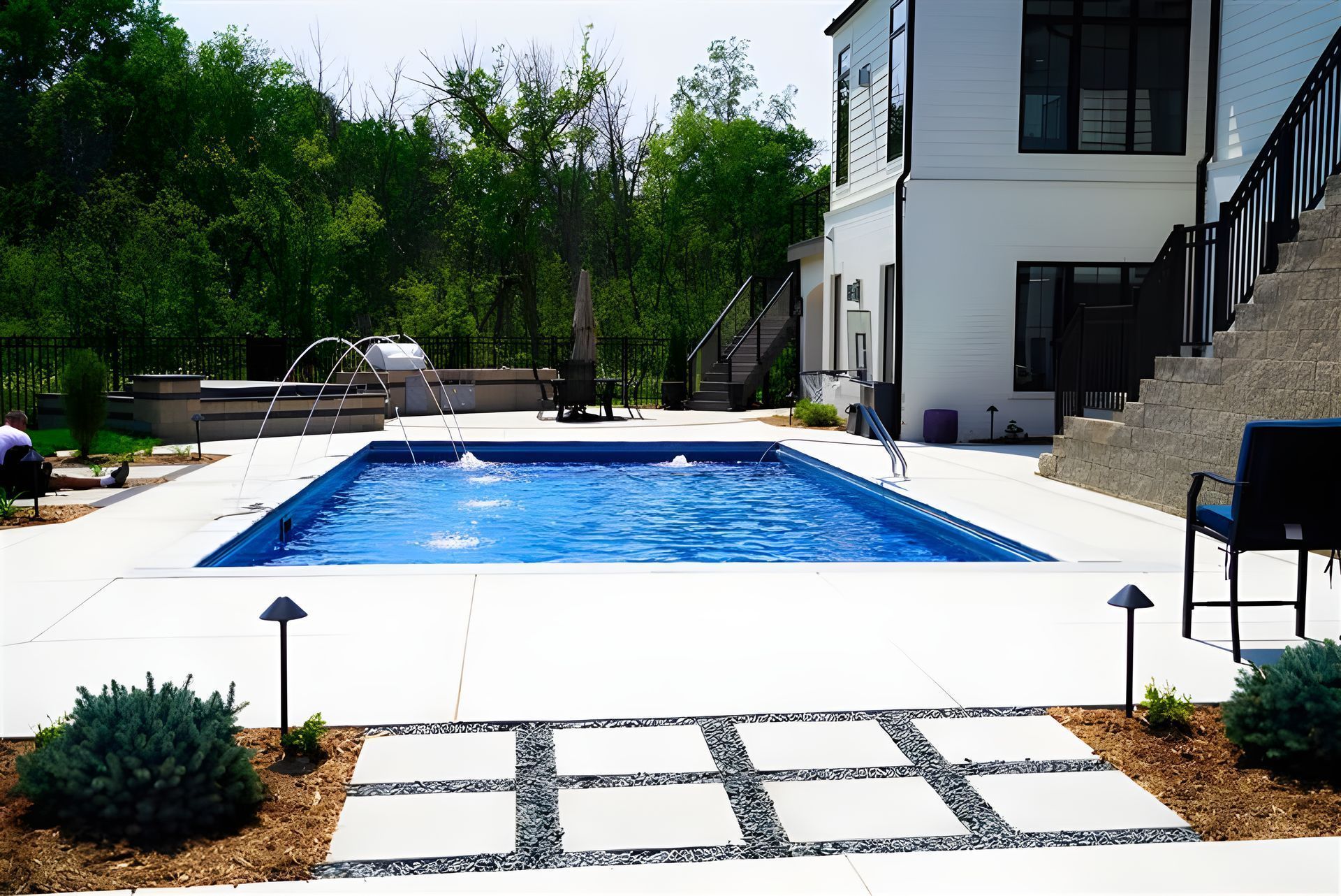 Pool in a backyard with stone pathway, white house, and trees. Blue water, sunny day.