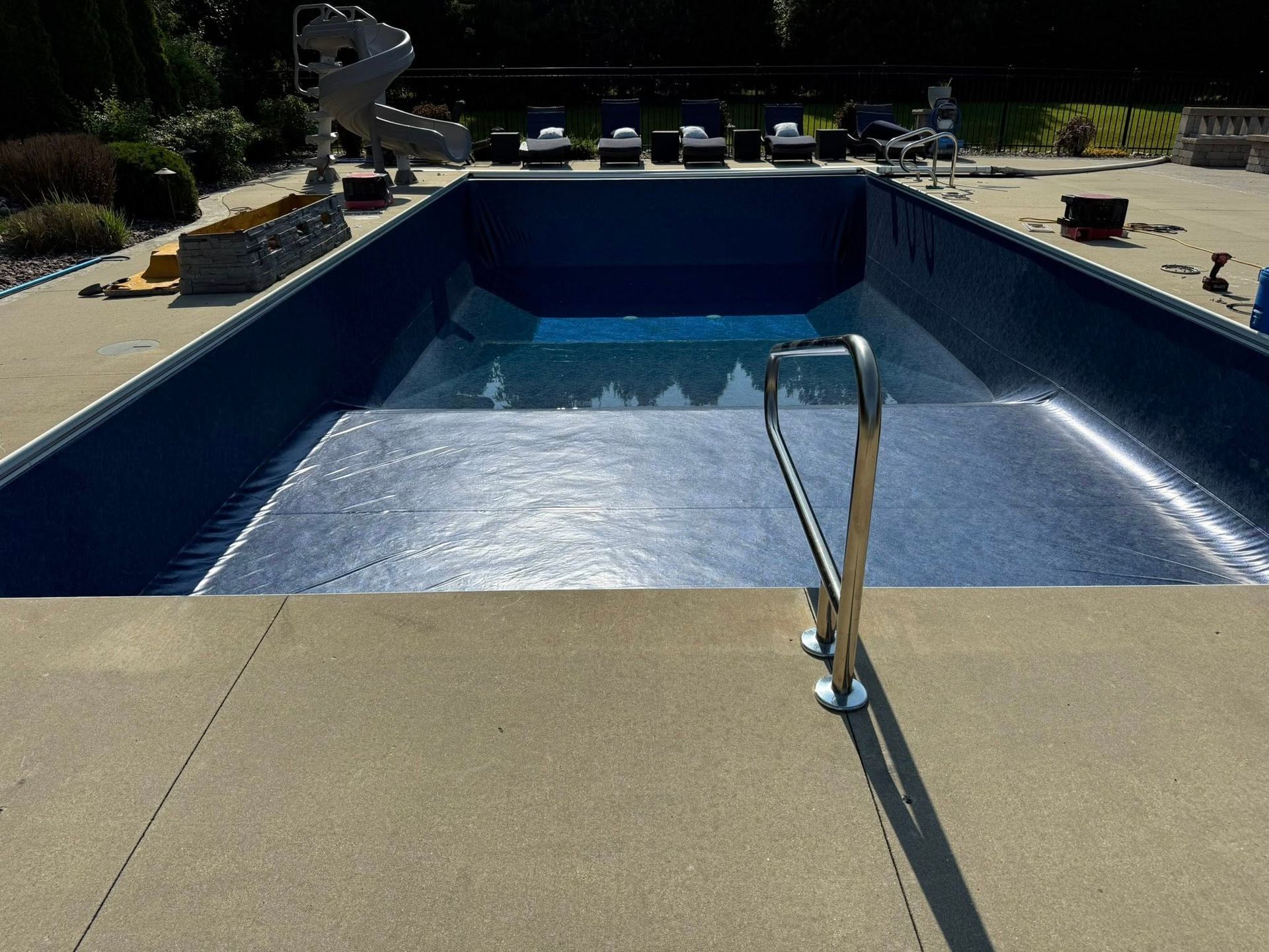 Empty blue-tiled swimming pool with steps, handrail, and concrete deck. Chairs and landscaping visible in background.