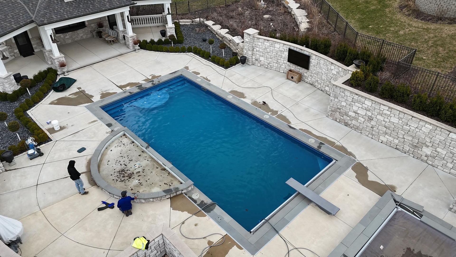 Aerial view of a rectangular blue swimming pool, patio, and stone walls. Two people are near the pool.