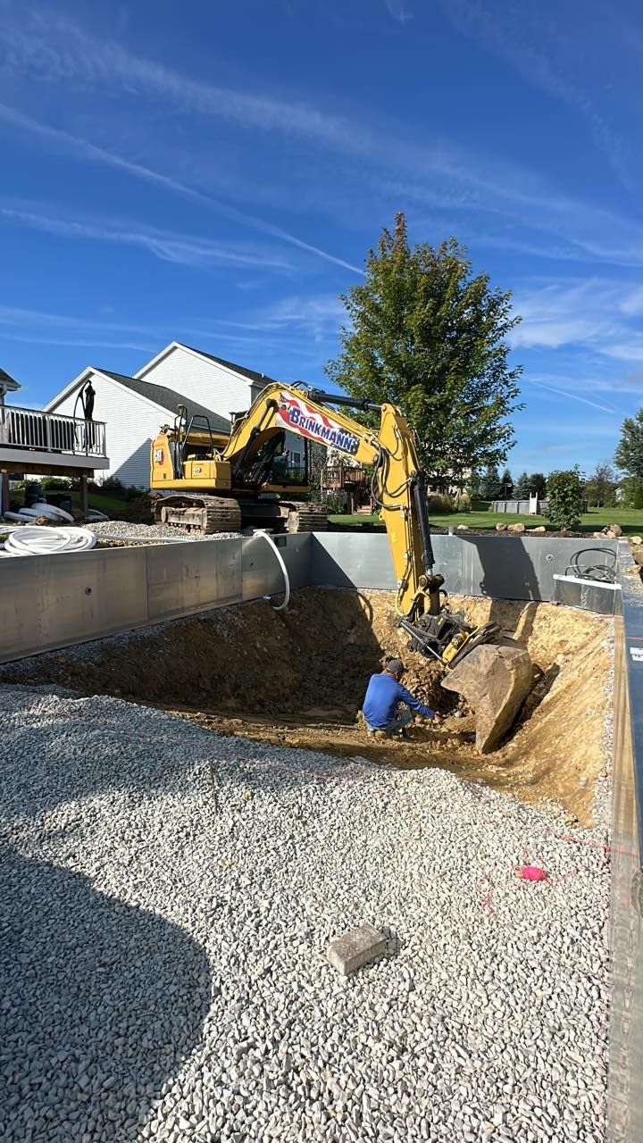 Excavator and worker in a construction site with gravel, a building, and a tree under a blue sky.