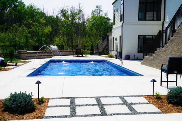 Rectangular blue pool surrounded by a concrete patio. Pathway with stone and squares leads to pool. House in background.