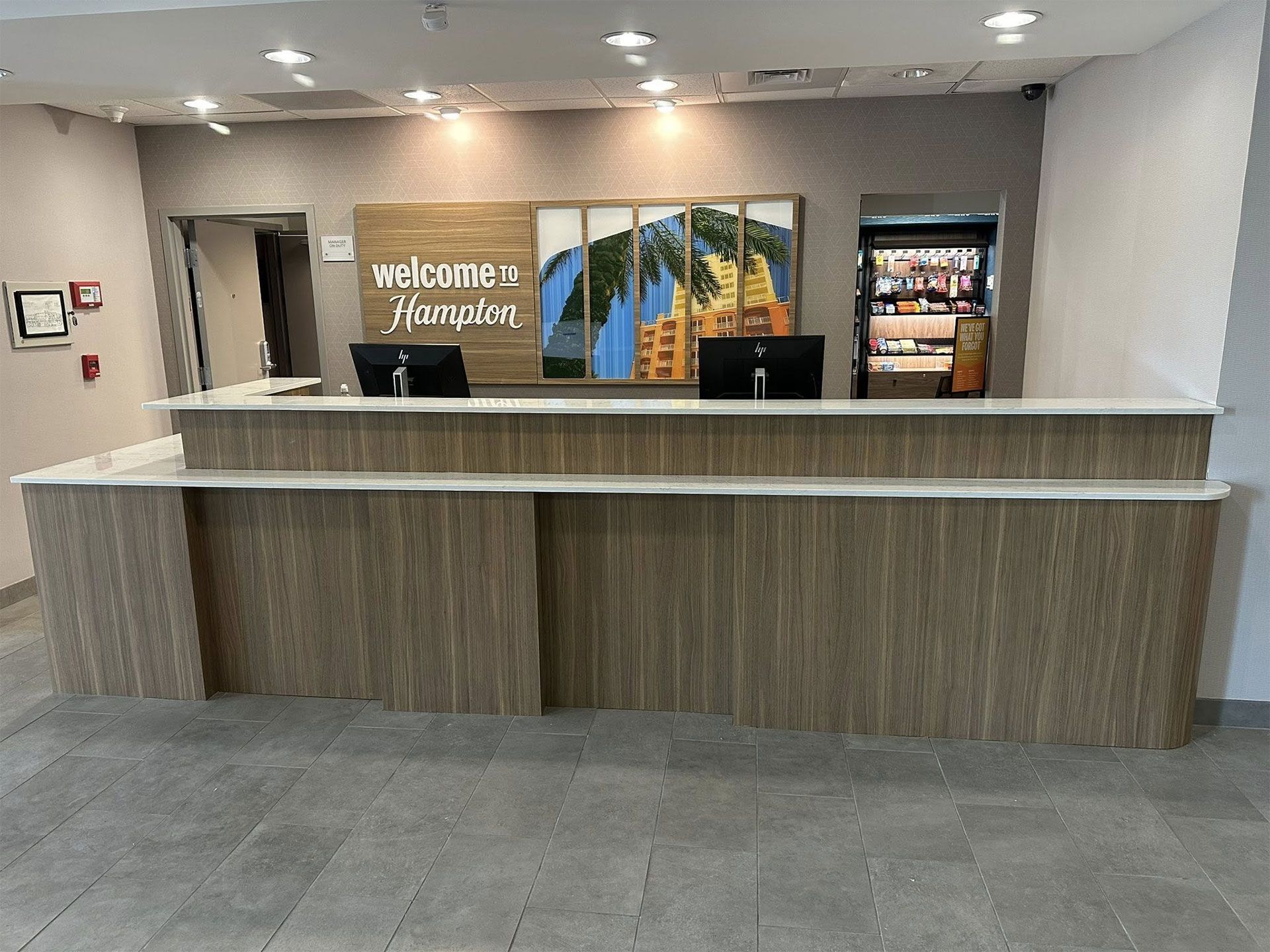 Hotel reception desk with "Welcome to Hampton" sign, computer monitors, and small store.