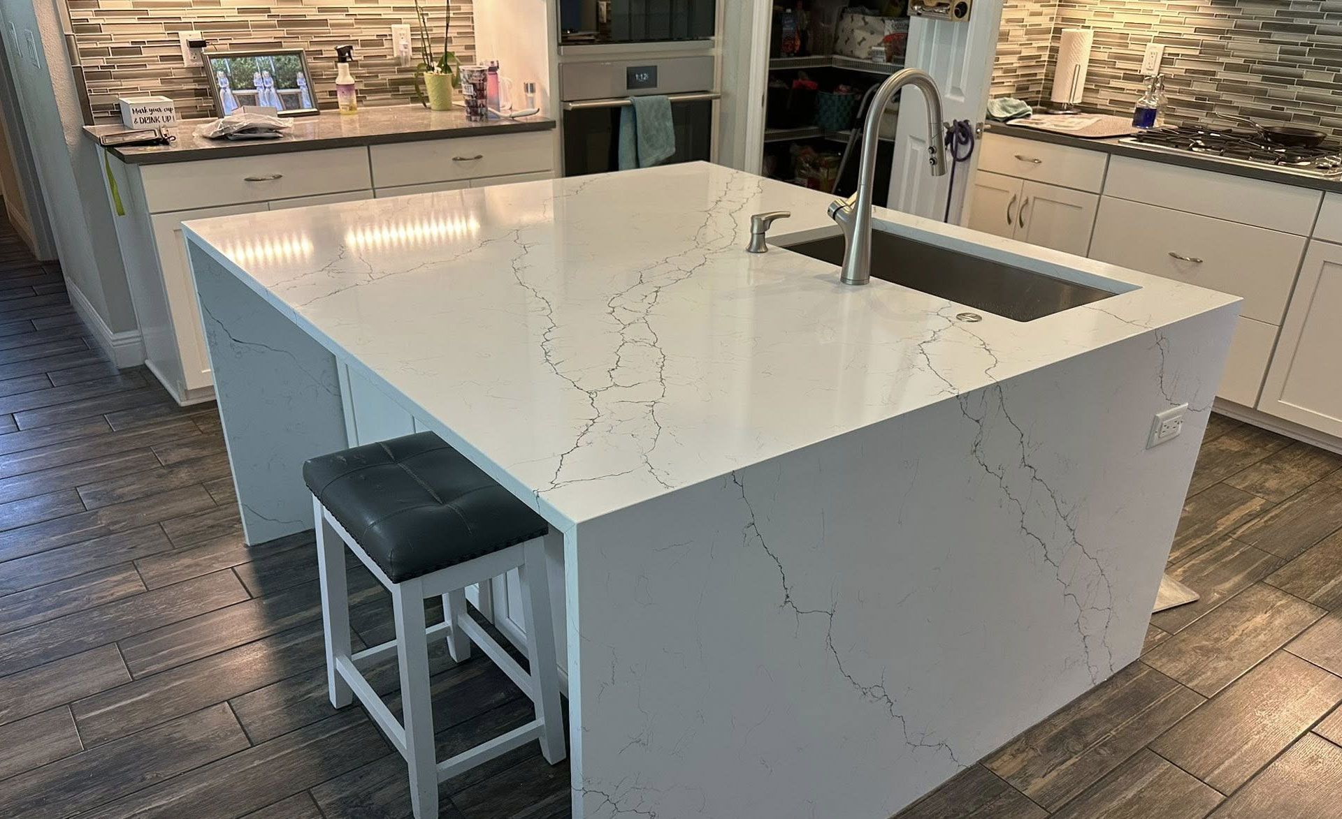 Kitchen island with white countertop, sink, and barstool. Wooden floor and cabinets in background.