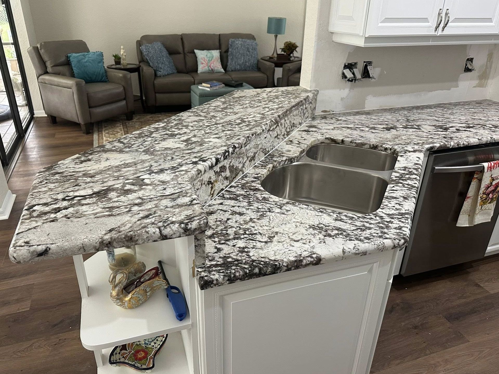 Kitchen island with granite countertop, double sink, white cabinets, and living room in the background.