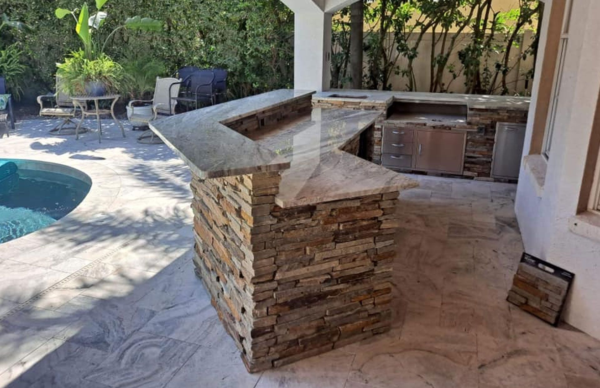 Outdoor kitchen with stone veneer and granite countertop, near a pool.