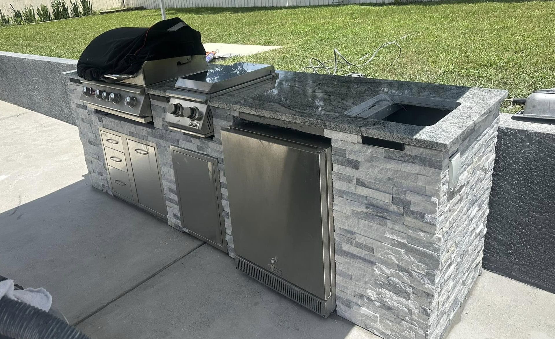 Outdoor kitchen with a grill, sink, and refrigerator, built into a gray stone structure.