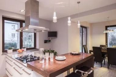 Modern kitchen with stainless steel range hood, island, dining area, and large windows.