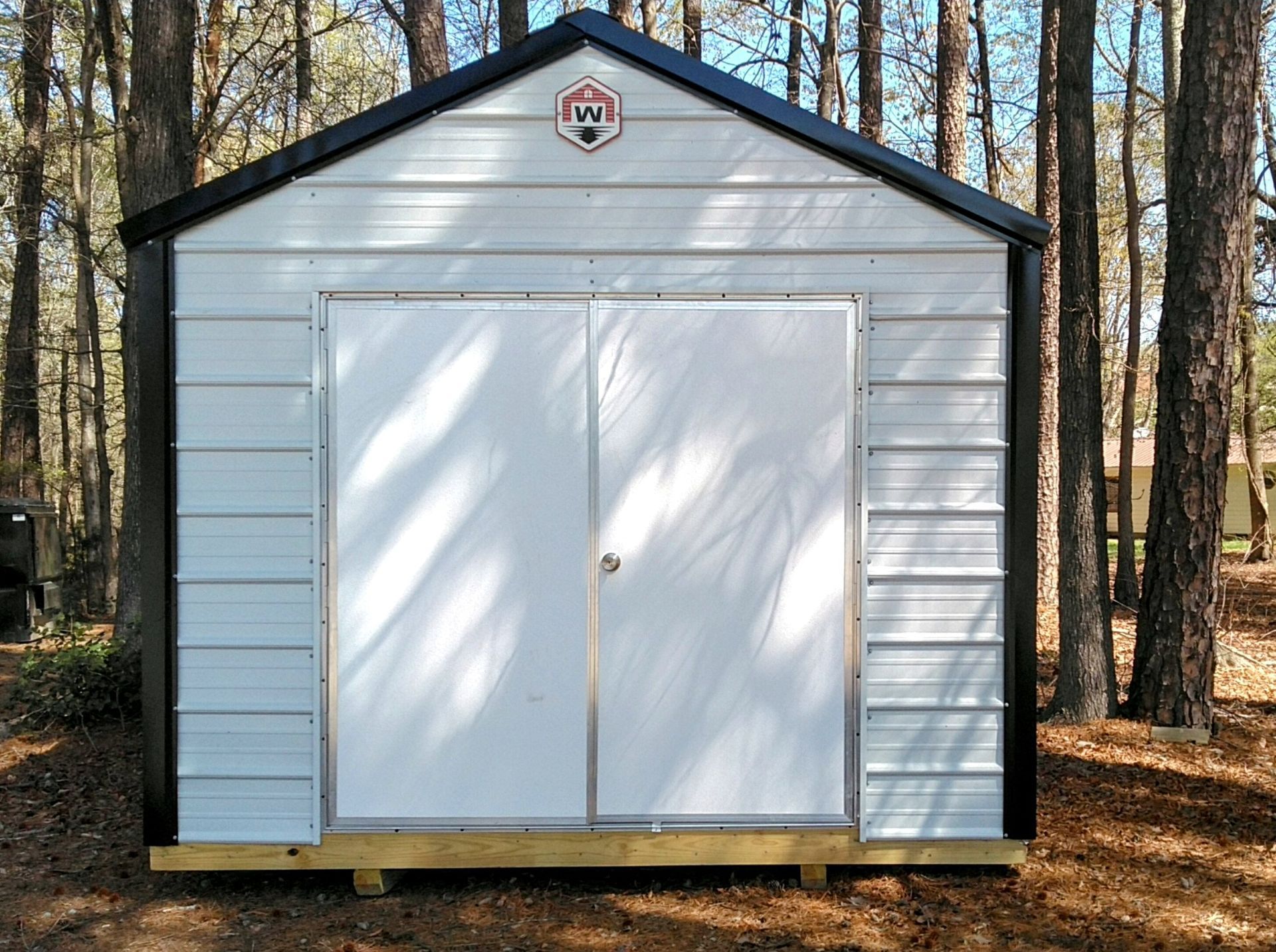 A white shed with black trim sits in the middle of a forest.