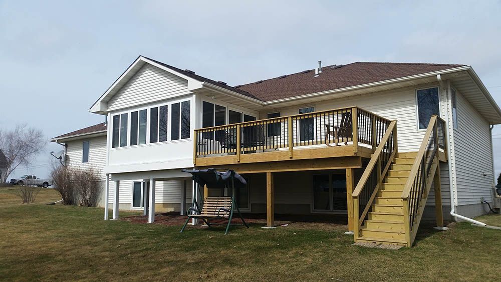 A two-story house with a wooden deck and stairs; brown roof, white siding, and green grass.