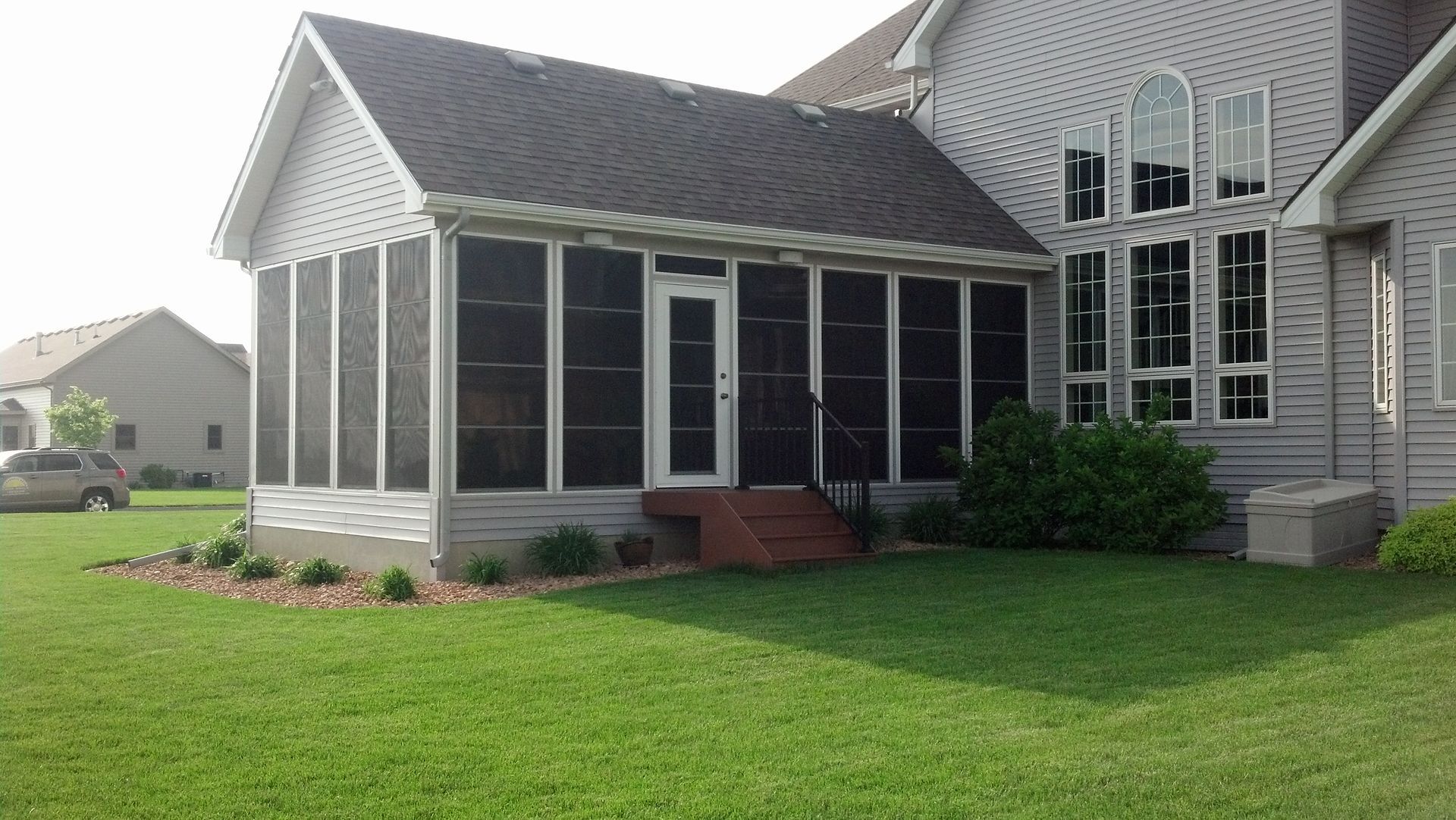 Screened porch with a door and steps attached to a house, on green grass, with a house in the background.