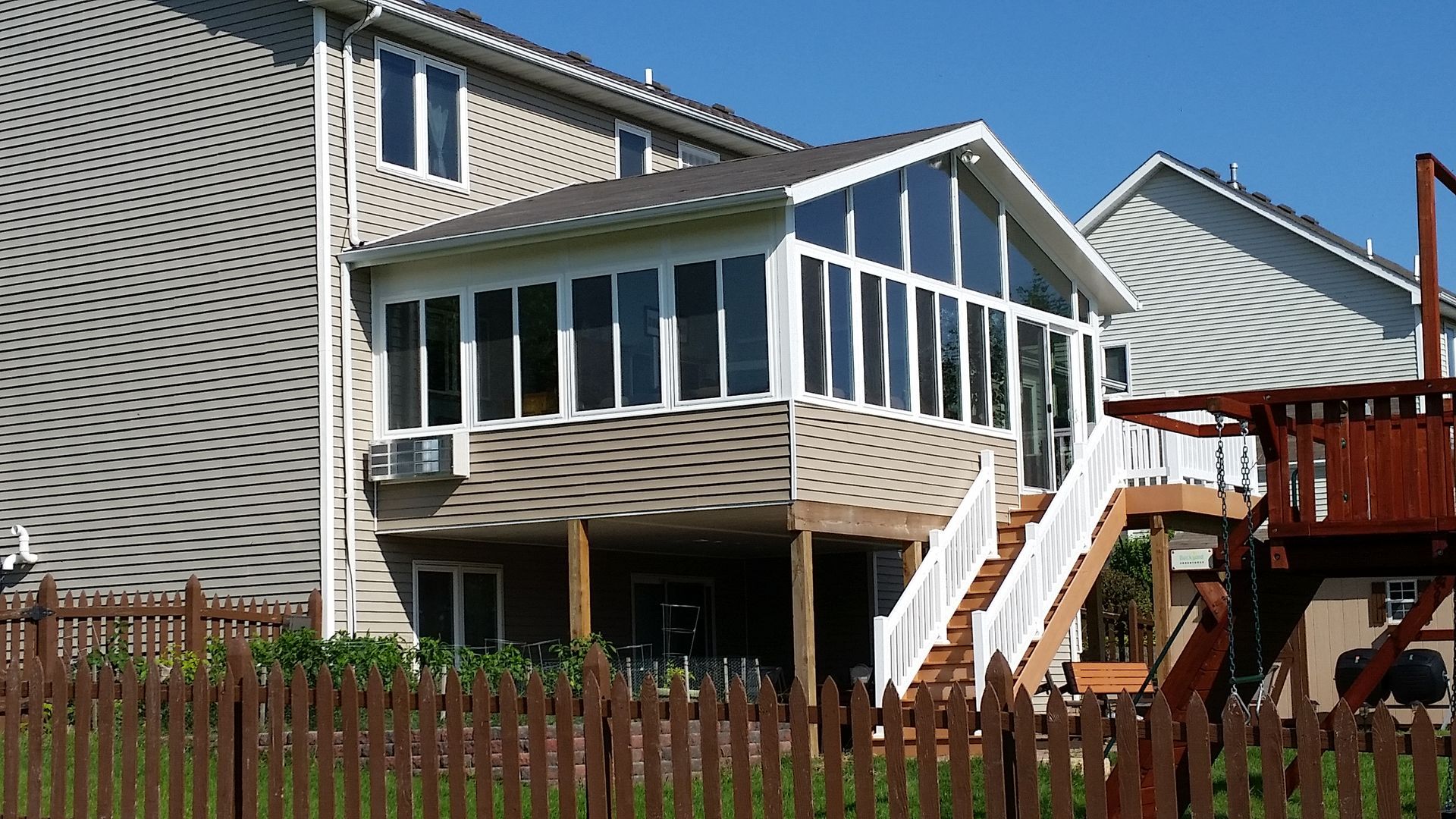 Screened-in porch attached to a two-story house with a deck and stairs, a brown picket fence, and a play set.